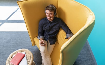 A man sits smiling in a yellow chair with a high back as he talks through a headset using his mobile phone