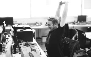 Happy man wearing black t-shirt stretches over his head at his desk in front of a computer in black and white