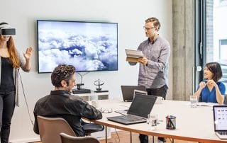 A meeting taking place while a young woman wears a virtual reality headset in from of an SEO flipboard while a man with a tablet looks on beside a tv screen showing clouds and others use their laptops