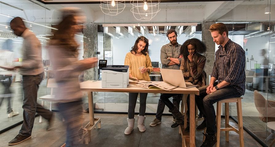 Desktop Xerox printer on wooden table with people looking at laptop, blurry woman in foreground, glass office