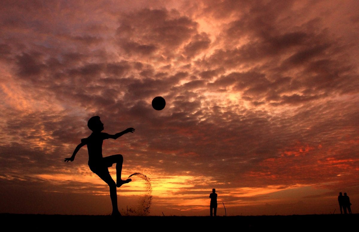 A silhouetted boy kicks a soccer ball in the foreground of a yellow, red, orange, and pink sunset and kicks up dirt