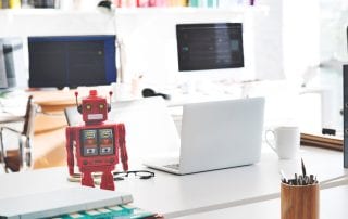 Red toy robot with yellow eyes on work desk with laptop, computer screens, books, pens, and papers
