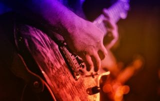 Hands close up playing an electric guitar under blue, purple, red, and orange stage lights