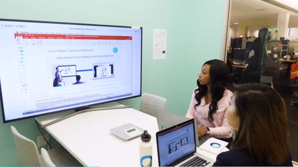 Two women sit at a desk in front of a Cisco webex monitor and laptop watching a business presentation